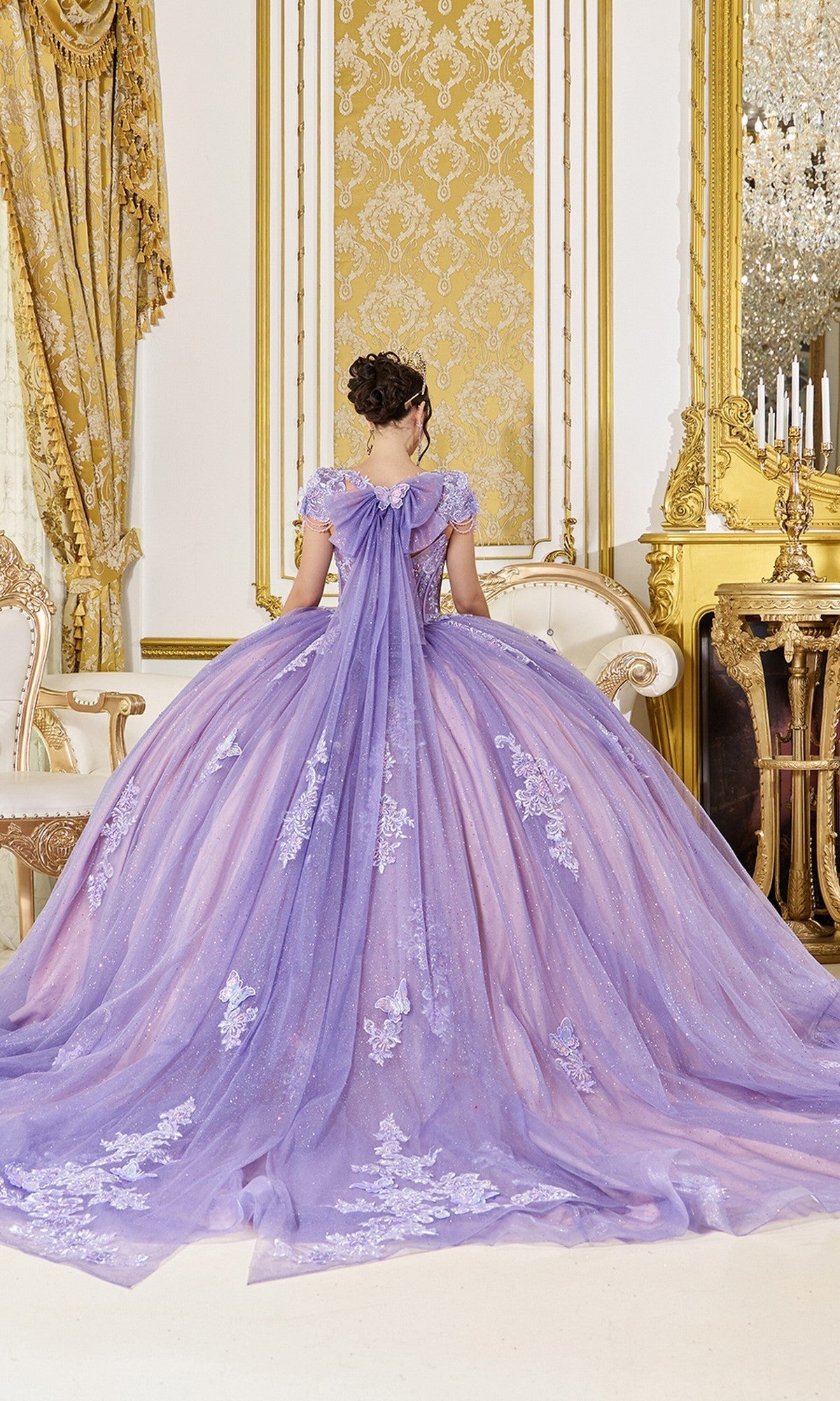 A woman in the Quinceanera Dress 5126 by Dancing Queen, a lavender ball gown with lace details and a corset bodice, stands facing away in an ornate room with gold accents and luxurious decor.